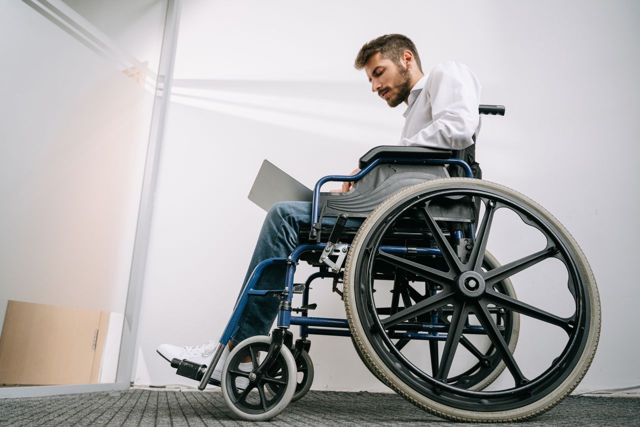 A man in a wheelchair concentrates on work using a laptop in an office setting.